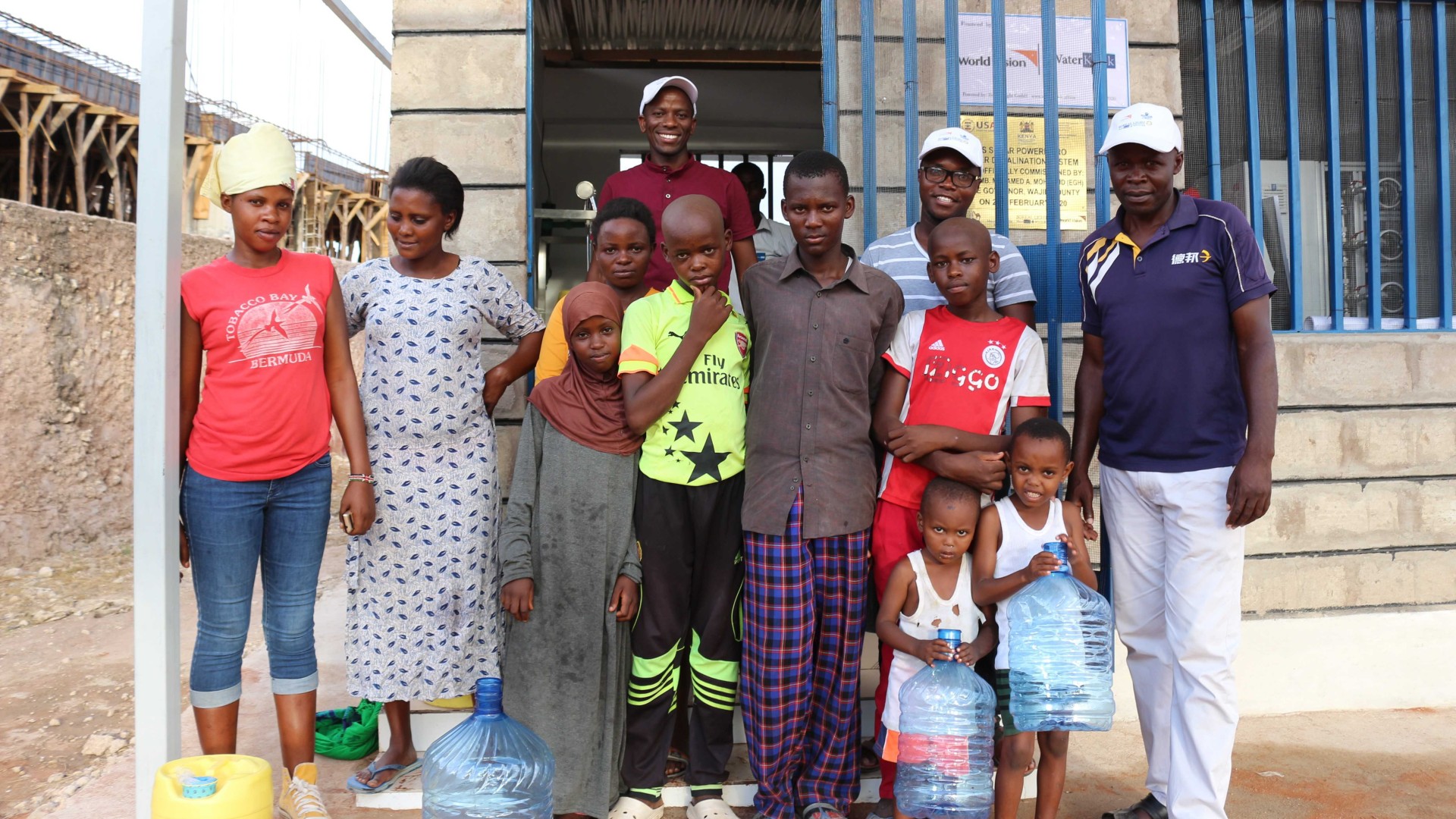 Younger and older villagers in Kenya stand with water canisters in front of a WaterKiosk