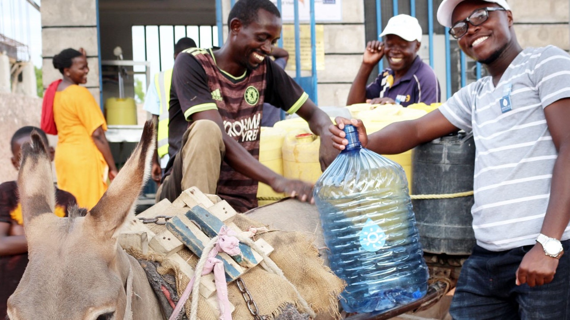 Two Kenyan young men stand laughing with a donkey in front of a WaterKiosk and attach a large bottle of drinking water to the donkey's back.