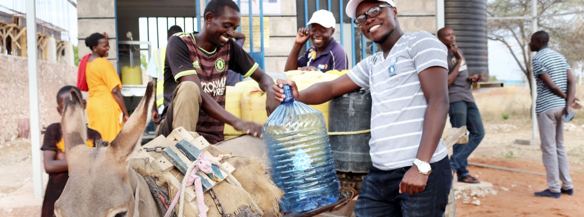 Two Kenyan young men stand laughing with a donkey in front of a WaterKiosk and attach a large bottle of drinking water to the donkey's back.