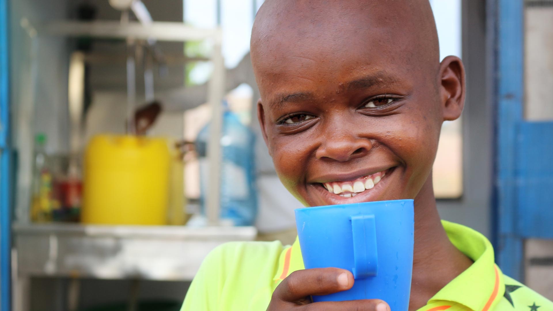 A Kenyan boy drinks water from a blue water cup and smiles.