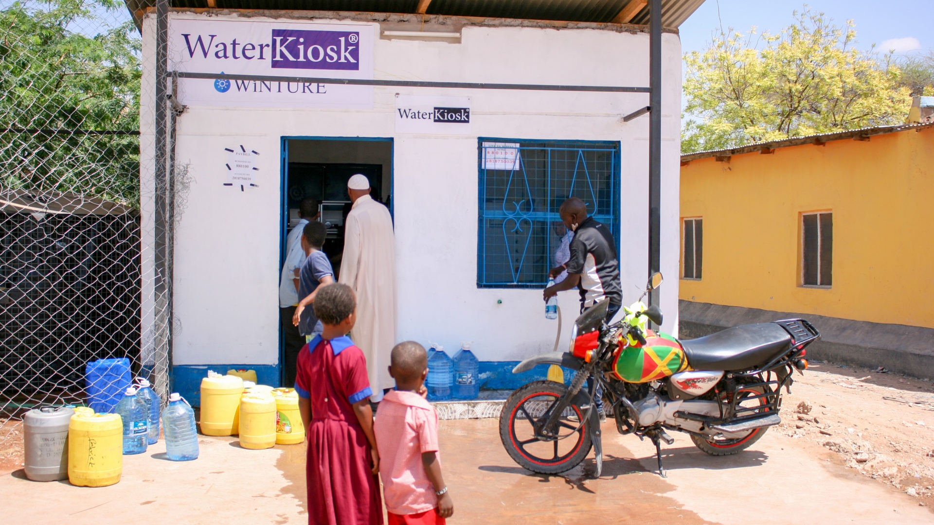 Ein Junger Kenianer füllt am WaterKiosk eine Falsche Trinkwasser ab. Sein Motorrad steht vor dem WaterKiosk und zwei kleine Kinder schauen zu.