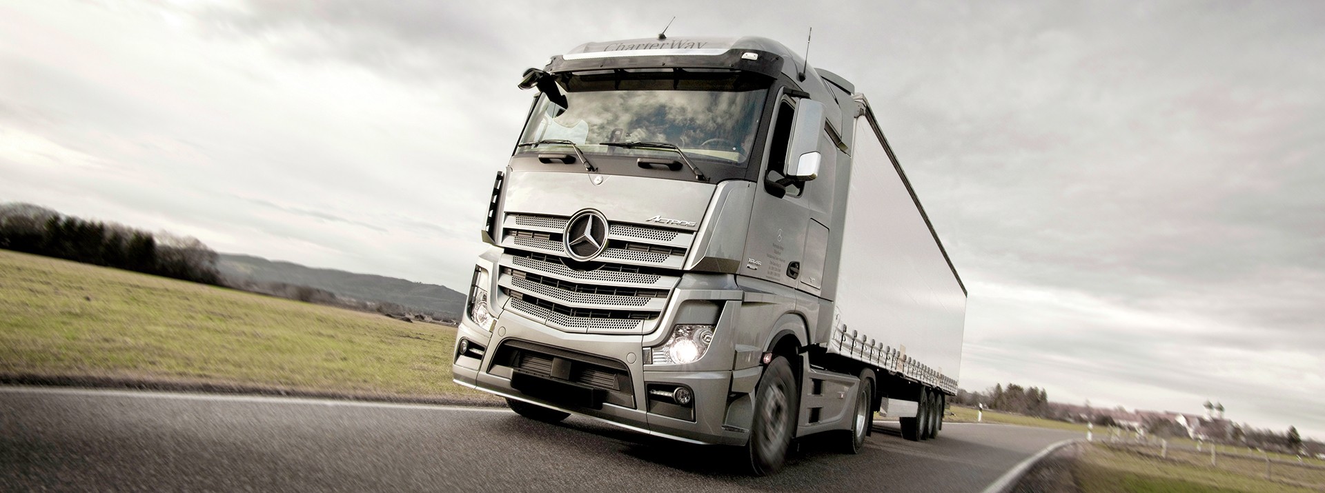 Moderner, silberner Lkw Actros von Meredes Benz fährt bei grauem, wolkenbehangenem Wetter auf einer Landstraße