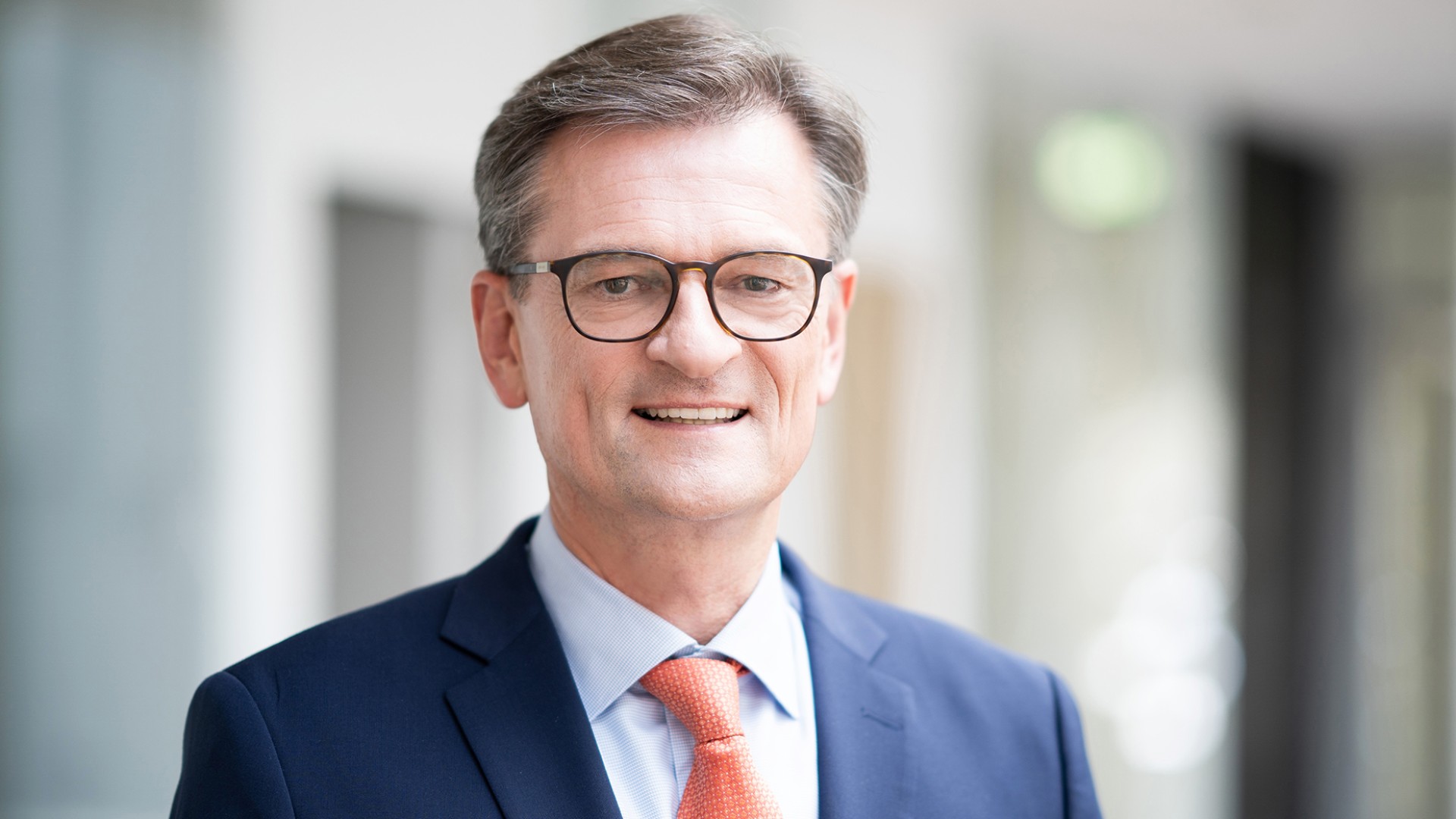 Portrait of Dr. Peter Radina, who is smiling in the photo. He wears dark brown glasses and a dark blue jacket with a light blue shirt and an orange-red patterned tie.