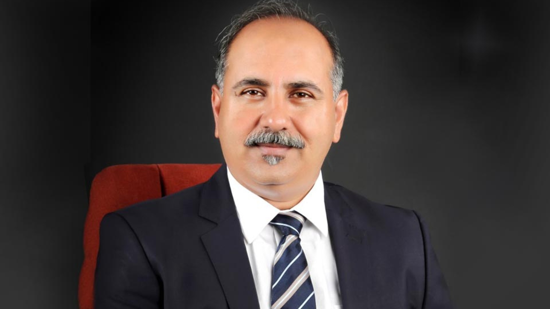 Portrait of Arun Kumar Bhat V S, Managing Director of Knorr-Bremse Technology Center India, showing him in a suit sitting on a red office chair against a dark background.