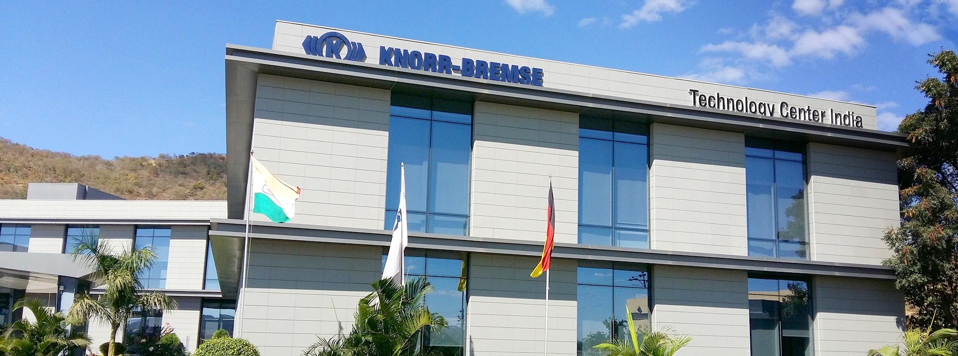 Technology Center India building at the Indian site in Pune. In the foreground palm trees and three flags, at the top of the building the Knorr-Bremse logo and the words "Technology Center India".
