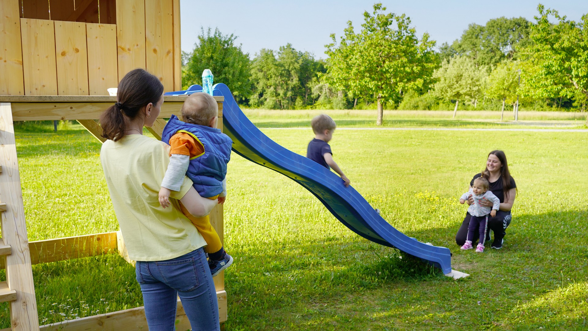 Zwei ukrainische Mütter spielen mit ihren Kleinkindern an einem Holzspielhaus im Garten des Hofgut Eck und lassen sie dort rutschen.