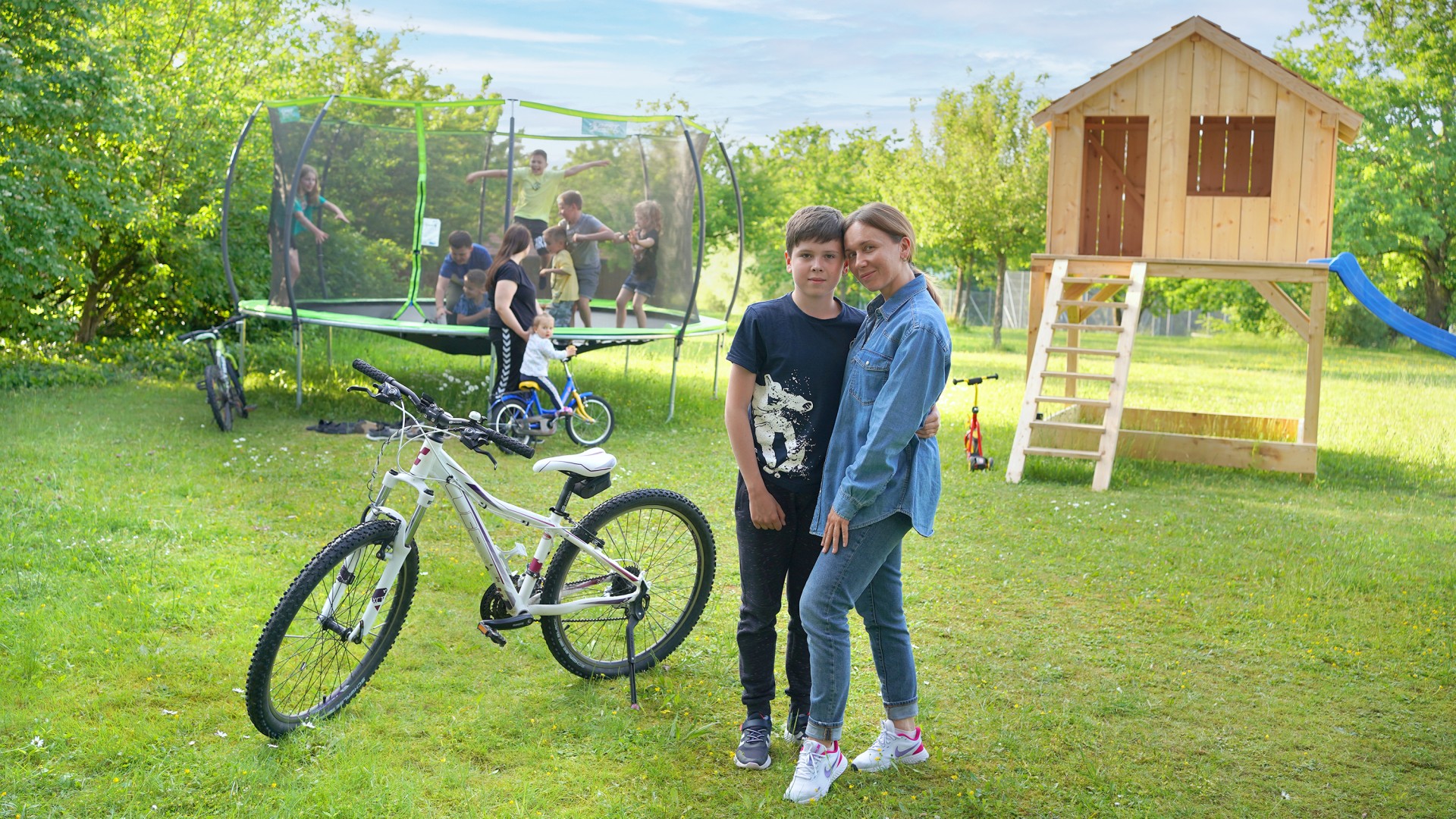 Eine ukrainische Mutter steht mit ihrem Jungen und einem Fahrrad vor einem Trampolin voller Kinder im Garten des Hofgut Eck.