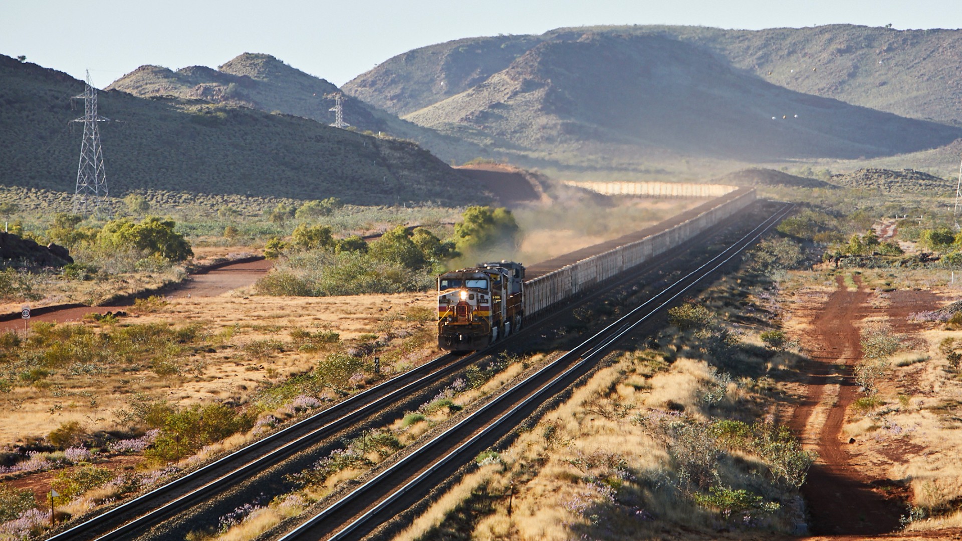 A 2.4 km long heavy freight train loaded with iron ore travels through a barren mountain landscape in Australia.