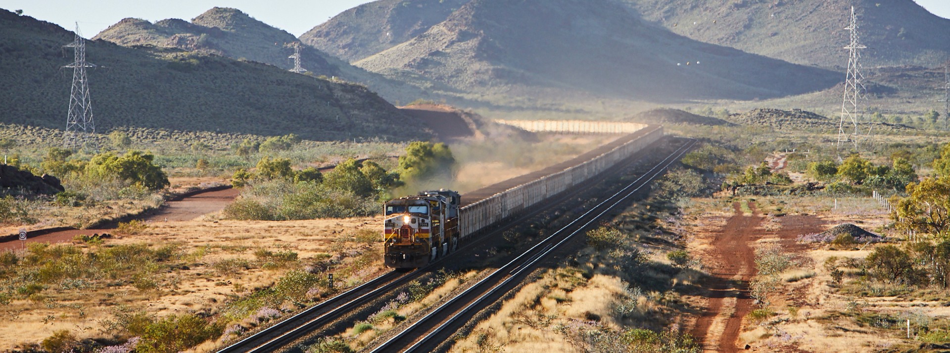 A 2.4 km long heavy freight train loaded with iron ore travels through a barren mountain landscape in Australia.