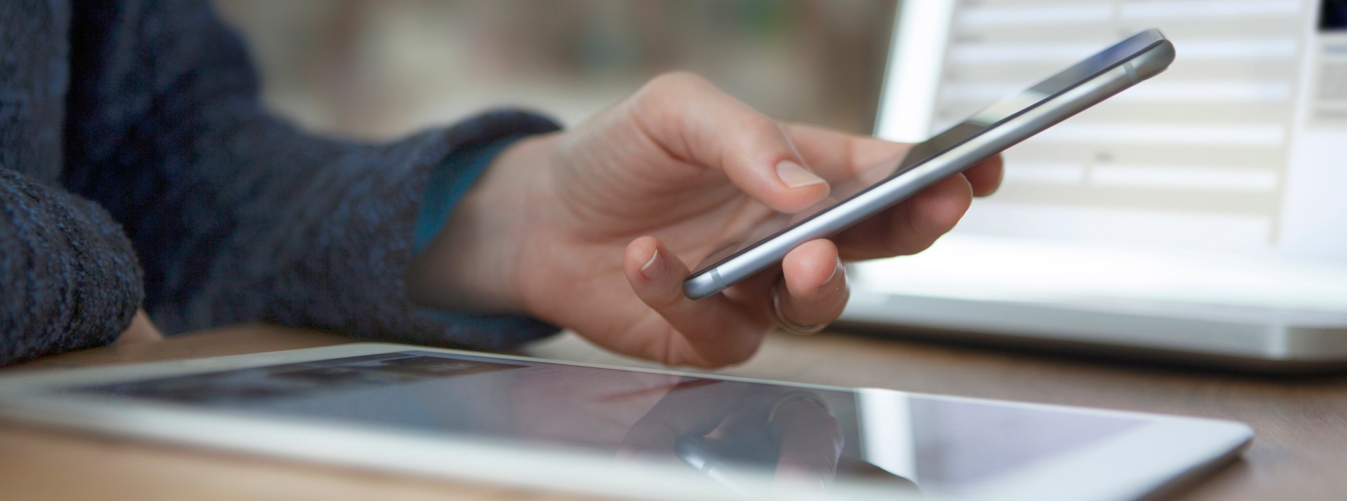 A hand in close-up holding a smartphone. In the background, an open laptop screen; in the foreground, a tablet lies on the table.