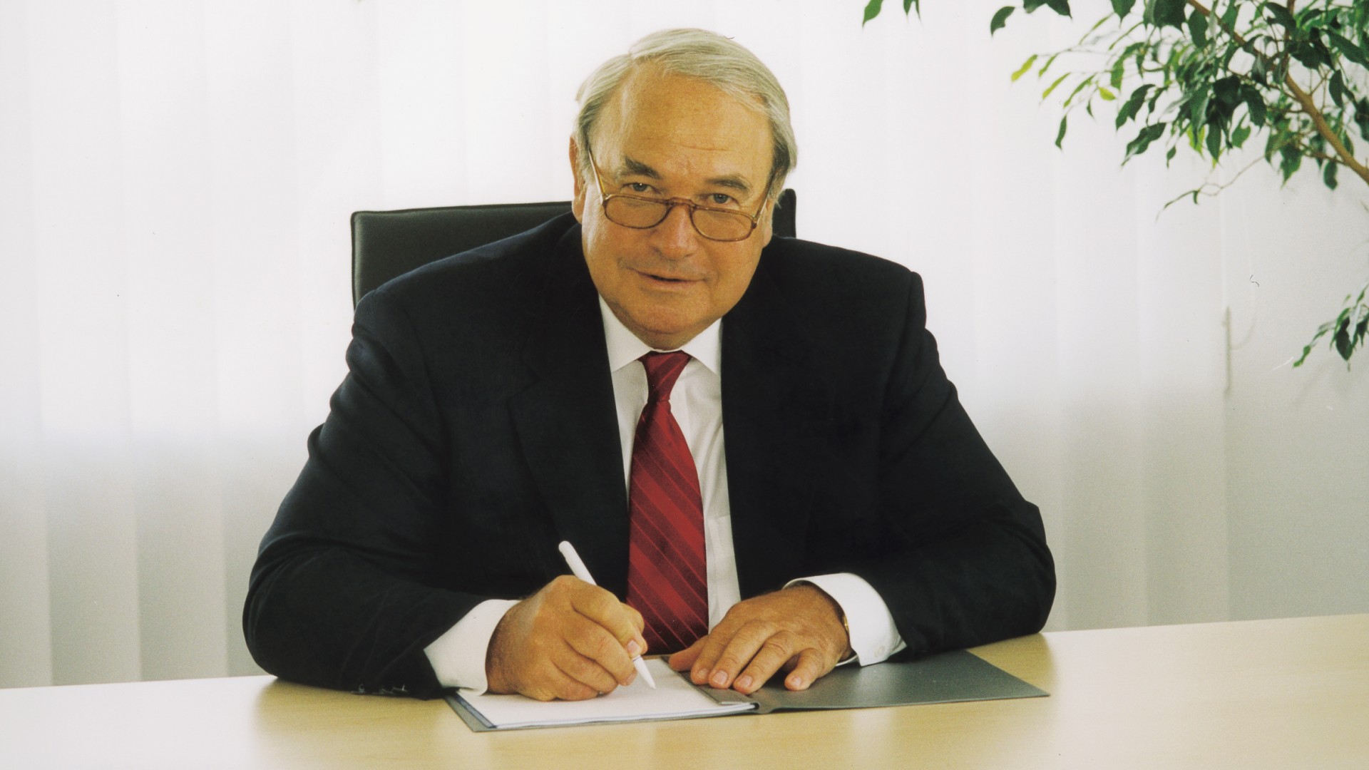 Person in a business suit signing a document at a desk in an office with natural daylight and a potted plant