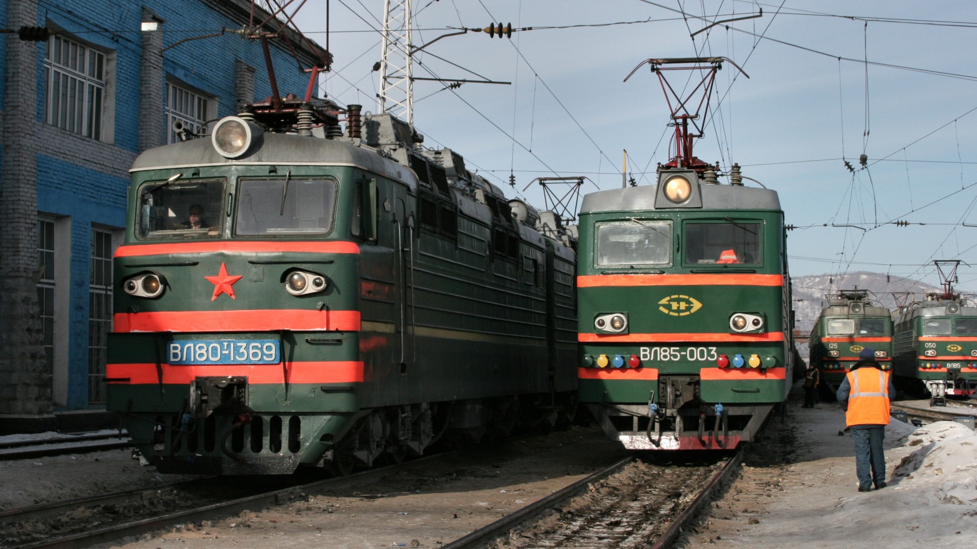 Grün‑rote Elektrolokomotiven auf einem verschneiten russischen Bahnbetriebsgelände mit Oberleitungen und einer Person in Warnweste