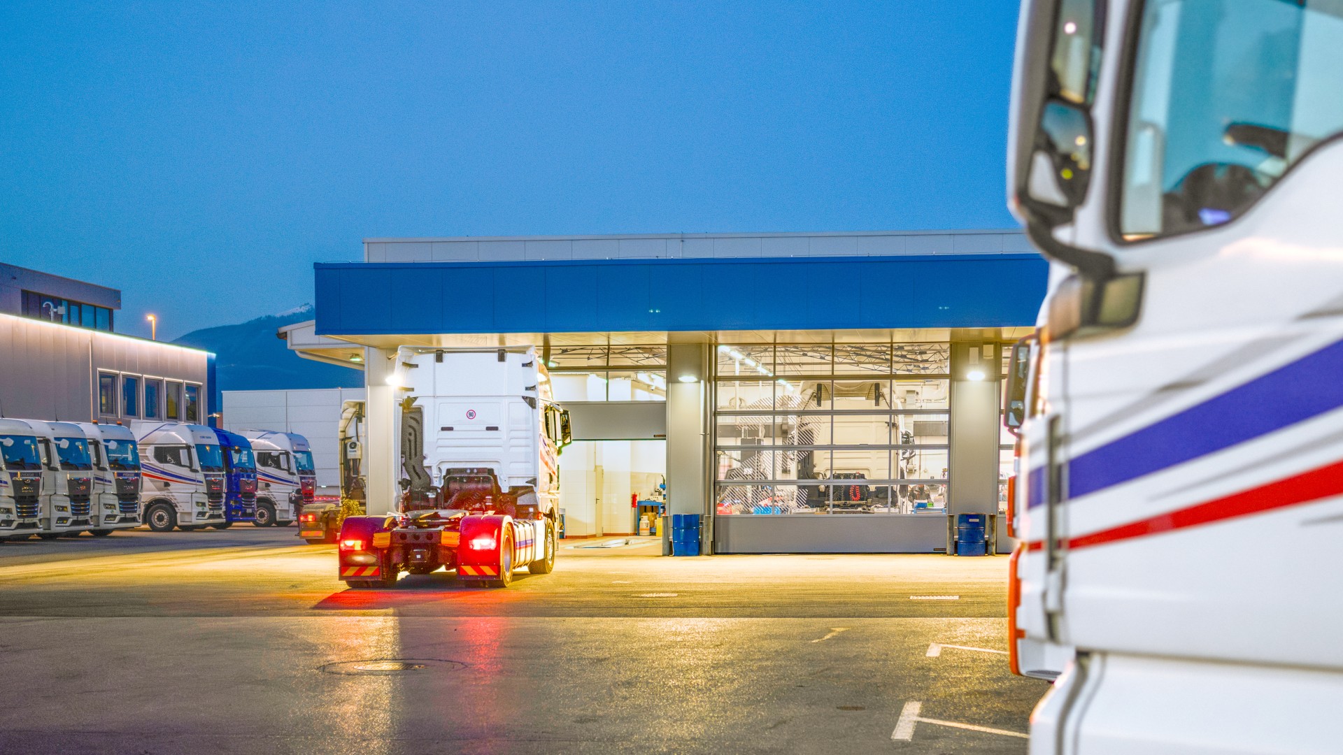 Nutzfahrzeuge vor einer beleuchteten Servicehalle einer Lkw‑Werkstatt bei Nacht