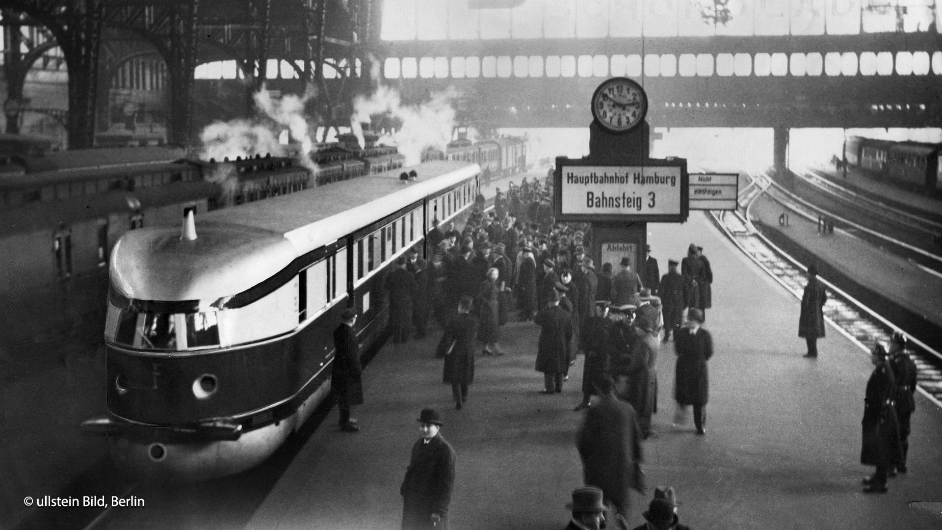 Historisches Schwarz-Weiß-Foto des Zugs ‚Fliegender Hamburger‘ bei der Einfahrt in den belebten Hamburger Hauptbahnhof.