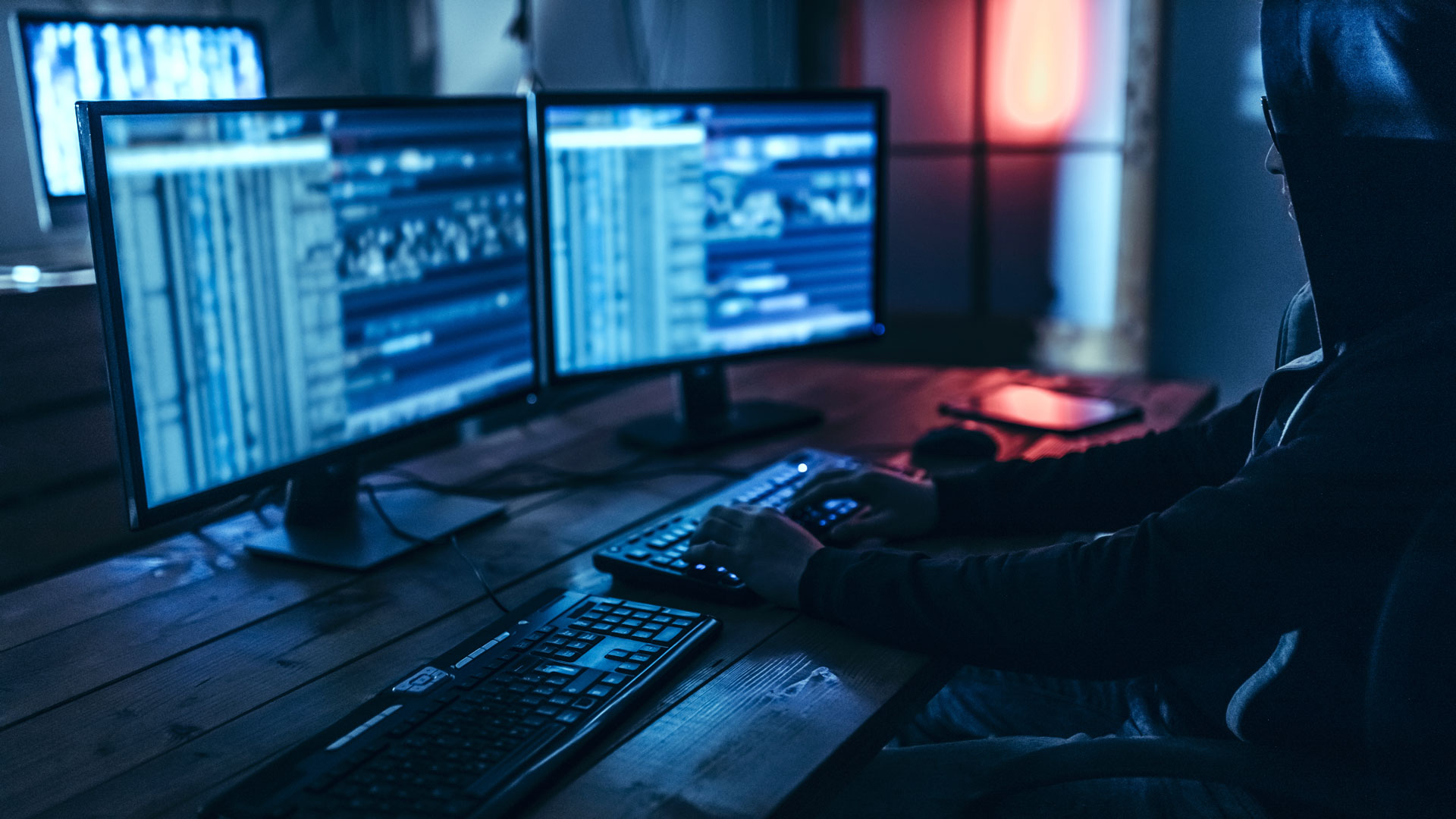 A young man in a hoodie, who is not recognizable, sits in a dark room in front of two screens and types on a keyboard