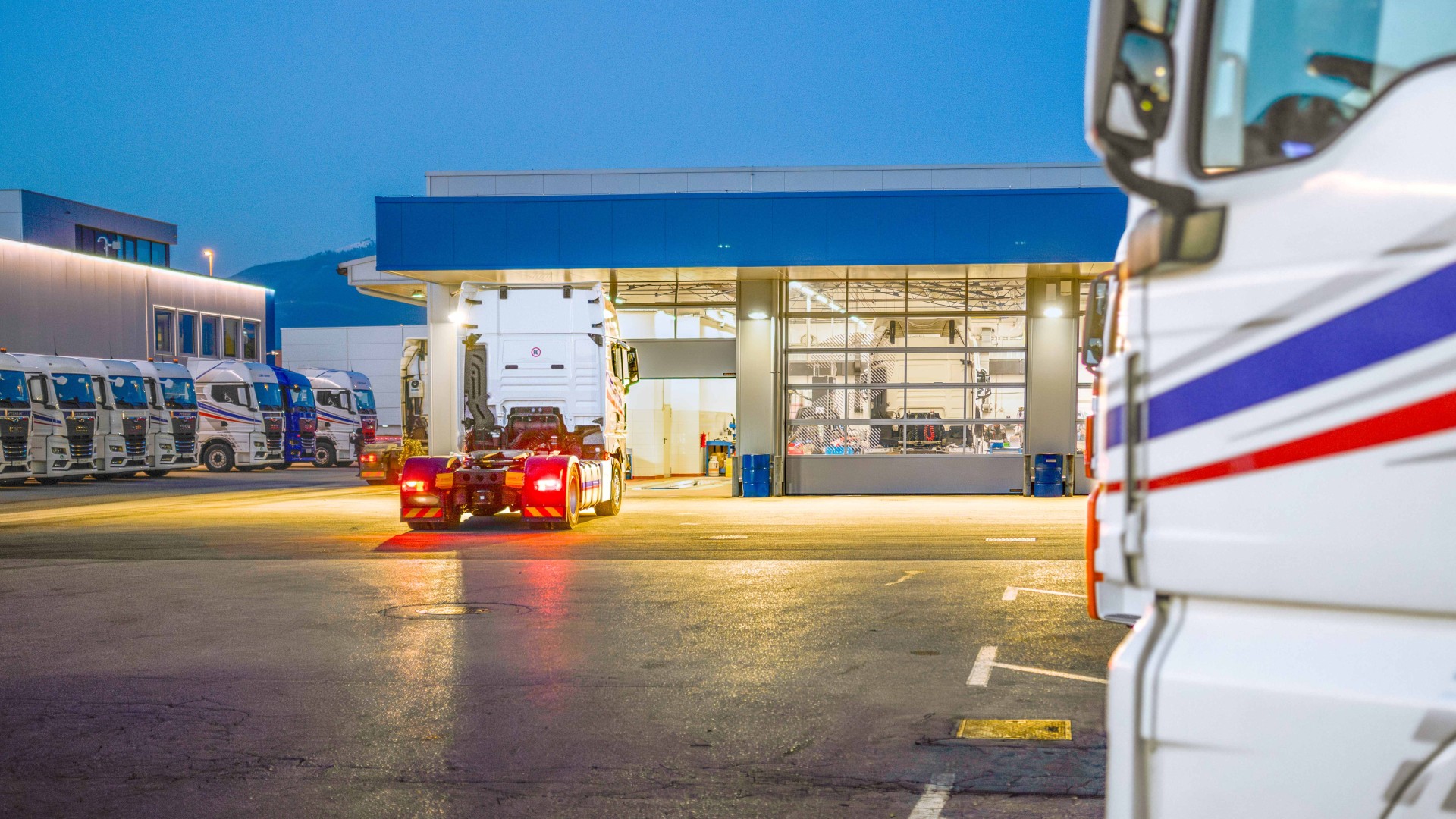 Lkw-Werkstatt bei Nacht: Eine Sattelzugmaschine ohne Auflieger fährt vor das Tor der beleuchteten Werkstatthalle, mehrere geparkte Lkw der Flotte stehen auf dem Speditionshof davor.