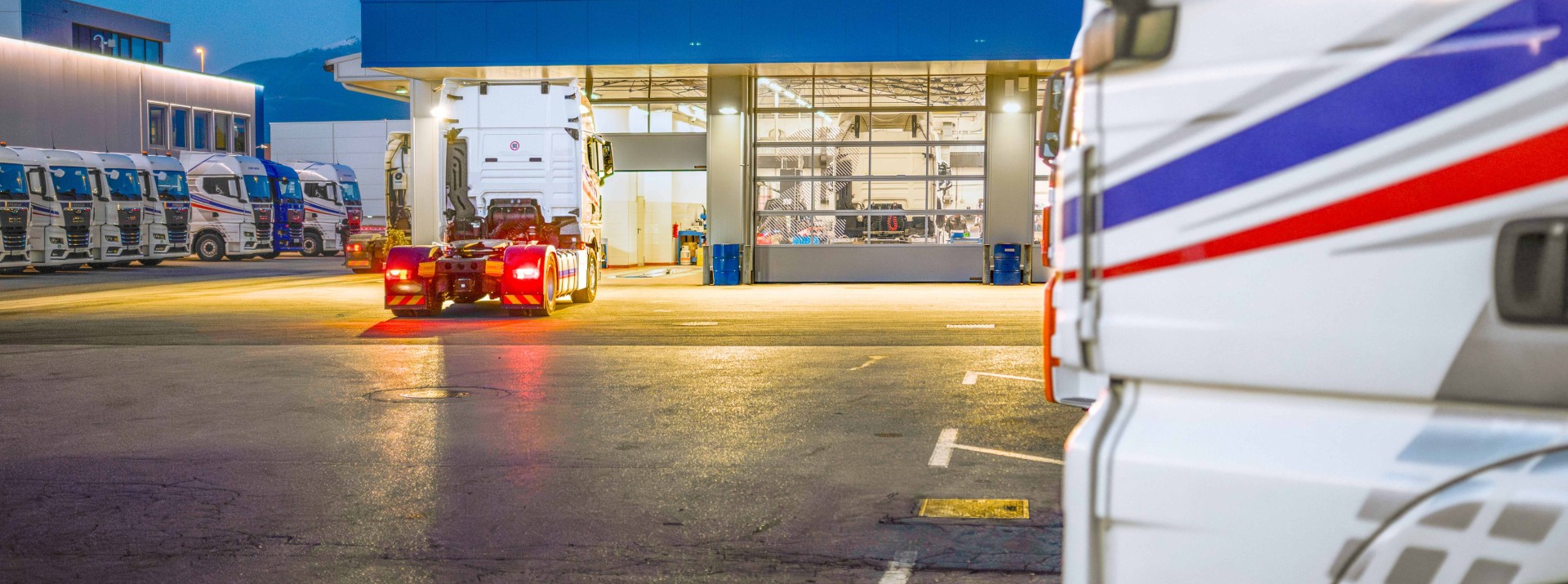 Lkw-Werkstatt bei Nacht: Eine Sattelzugmaschine ohne Auflieger fährt vor das Tor der beleuchteten Werkstatthalle, mehrere geparkte Lkw der Flotte stehen auf dem Speditionshof davor.