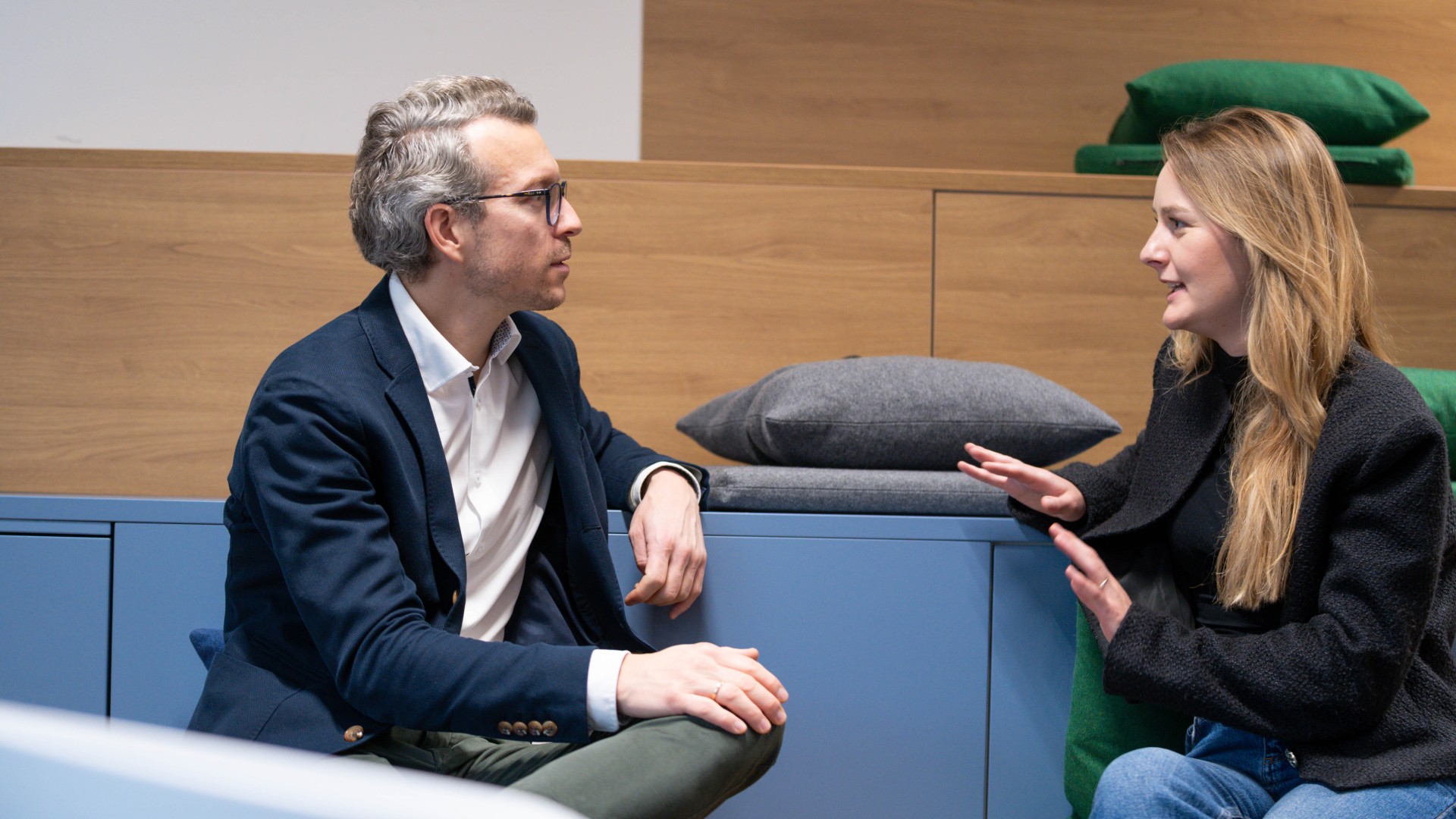 Two employees engaged in a reverse‑mentoring conversation in a modern lounge setting with seating steps and cushions