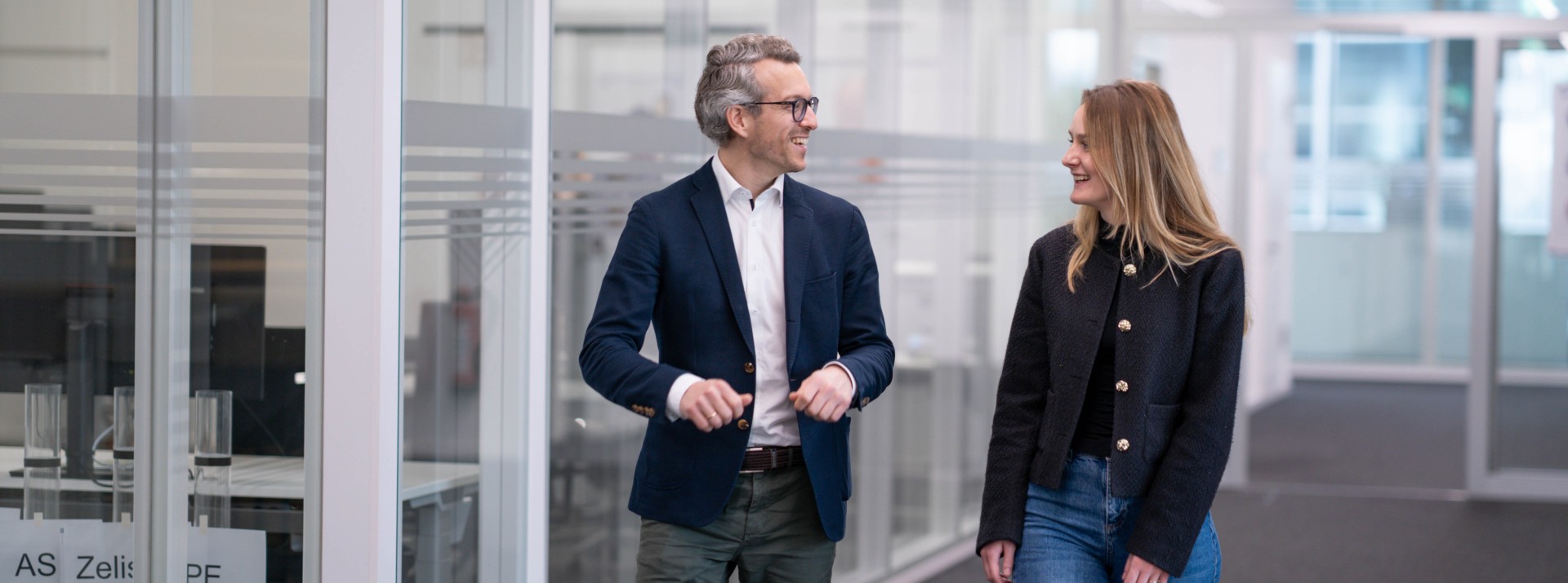 Two employees engaged in a reverse‑mentoring exchange while walking through a modern office corridor with glass walls
