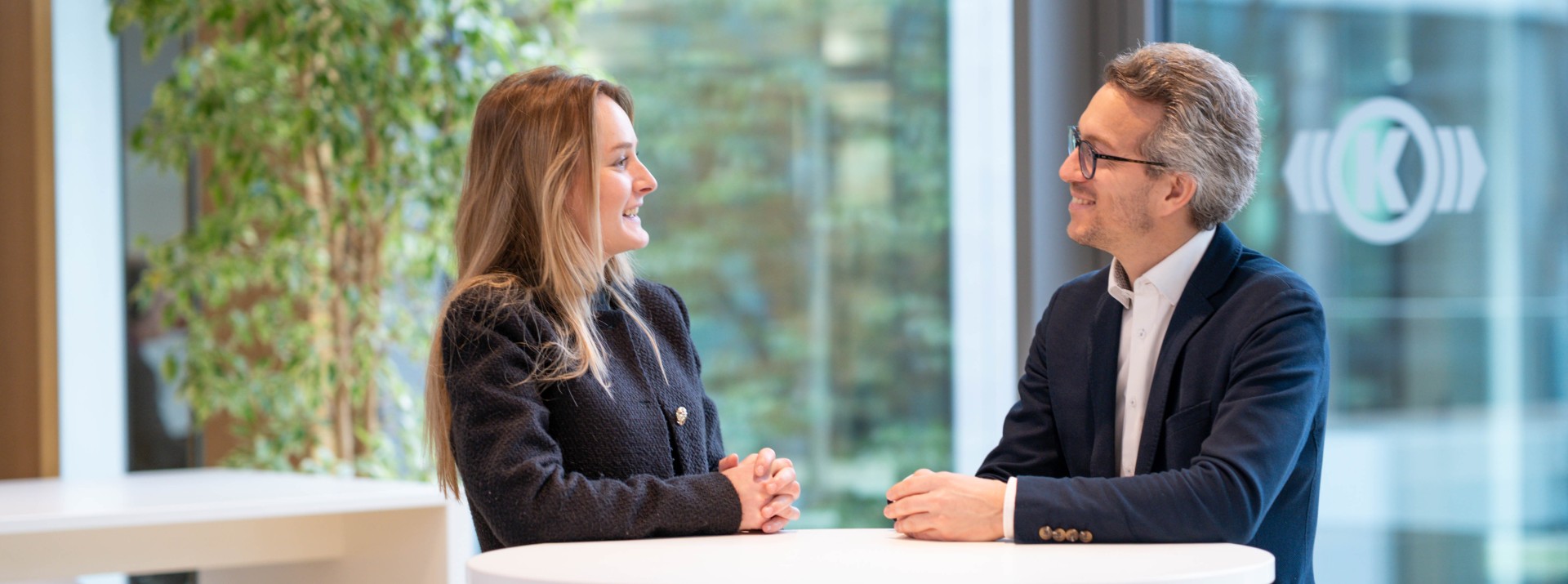 Two employees engaged in a reverse‑mentoring exchange at a standing table in a modern office lounge area with large windows and plants.