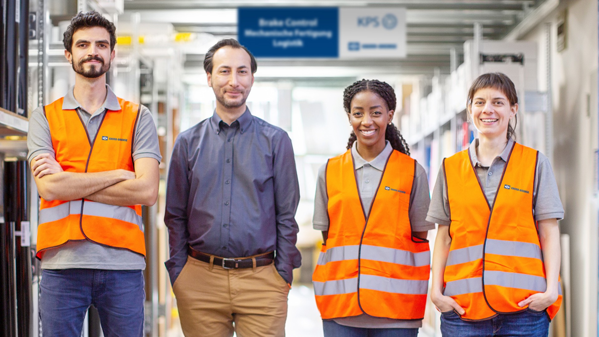 Four Knorr‑Bremse employees standing in a production hall; the diverse team, some wearing safety vests, is positioned between shelves in the manufacturing and logistics area